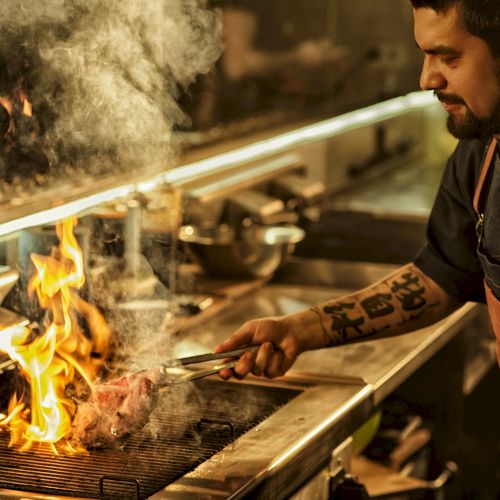 A chef in a kitchen grills over an open flame, flames licking a piece of meat as smoke rises, focused on cooking in a busy station.