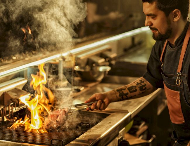 A chef in a kitchen grills over an open flame, flames licking a piece of meat as smoke rises, focused on cooking in a busy station.